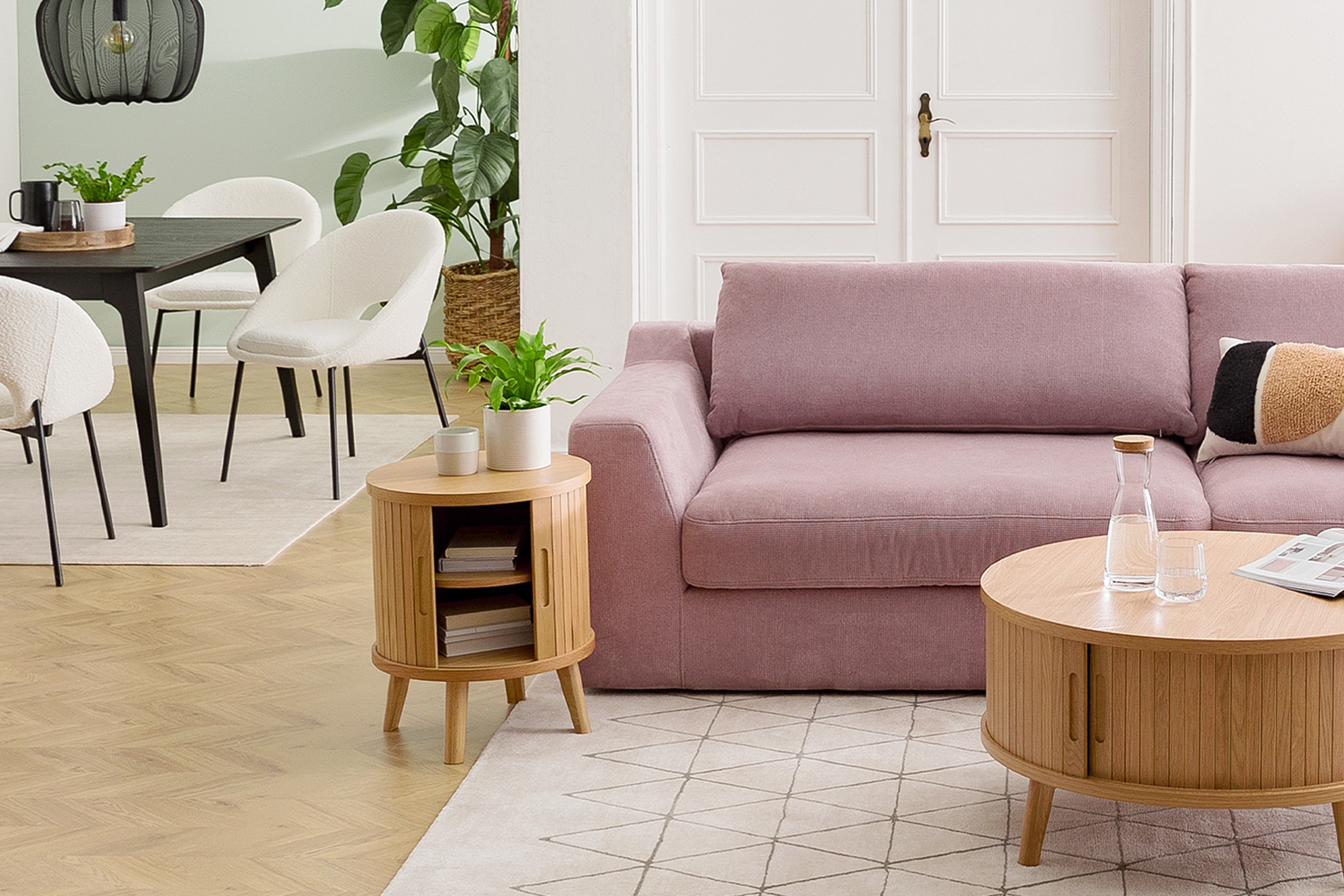 Offener Wohn- und Essbereich mit dem Trend-Bigsofa Dixwell von home24 als Ankerpunkt, dazu Stauraummöbel aus hellem Holz im Retro-Stil sowie ein schwarzer Esstisch, schwarz-weiße Esszimmerstühle, Teppiche, schwarze Lampen und Grünpflanzen.