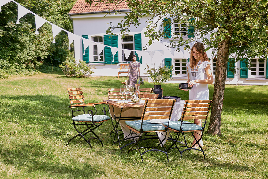 Klassische Holzklappmöbel inmitten eines sonnigen, grünen Gartens mit geschwungenen Lehnen, weißer Wimpelkette über dem Tisch und zwei Frauen, die den Tisch decken