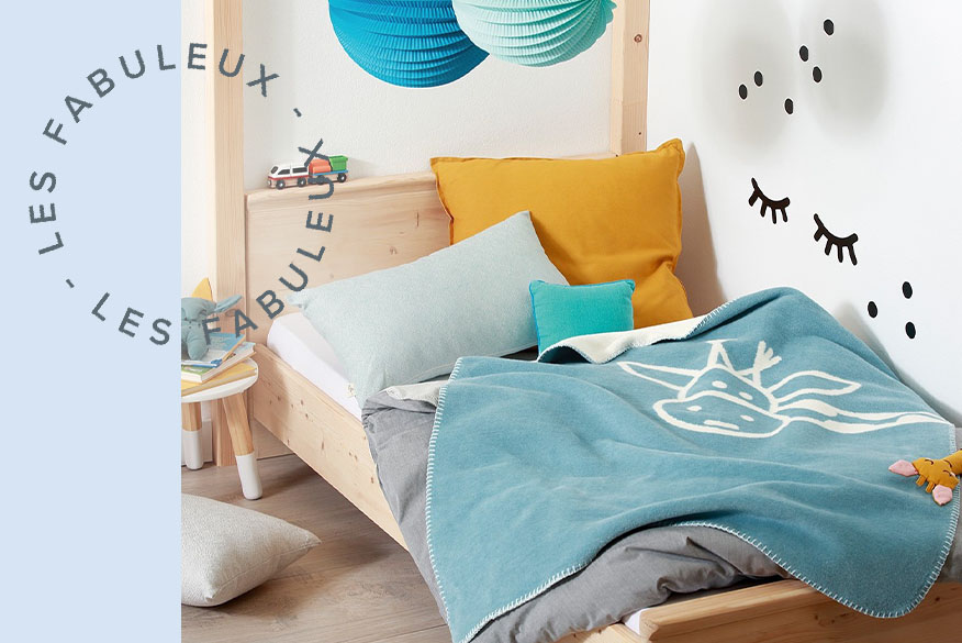 Lit enfant en bois blond avec plaids et coussins en ocre jaune, bleu lagon et gris pierre ; tapis rond beige à franges, coussin avec arc-en-ciel et jeu premier âge