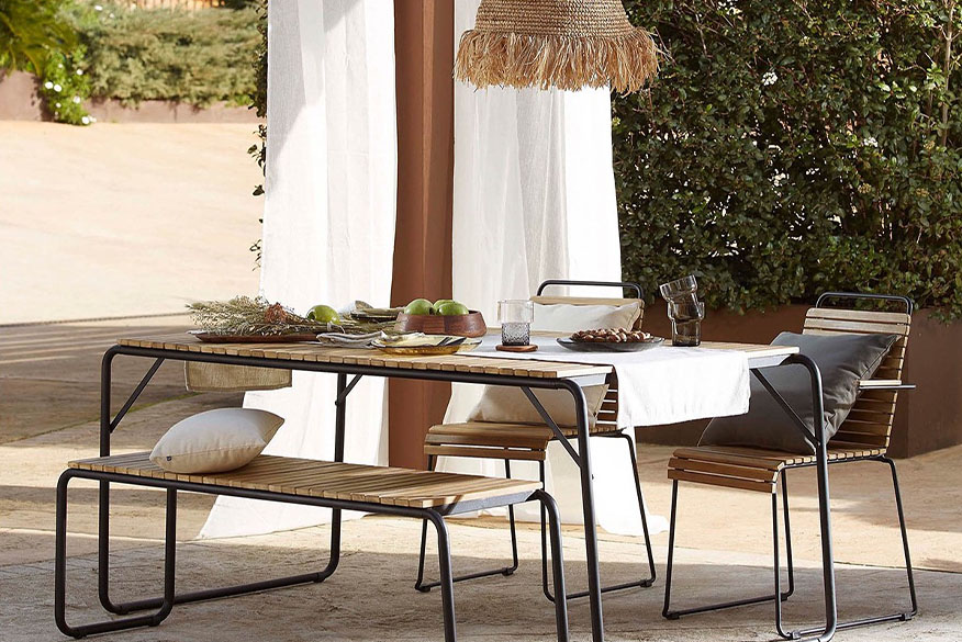 Terrasse in mediterranem Finca-Style mit Gartemöbeln aus Akazienholz und Stahlgestell, die kombiniert sind mit cremefarbenen Outdoor-Textilien, einer Pendellampe in Stroh-Optik und weißen Vorhängen