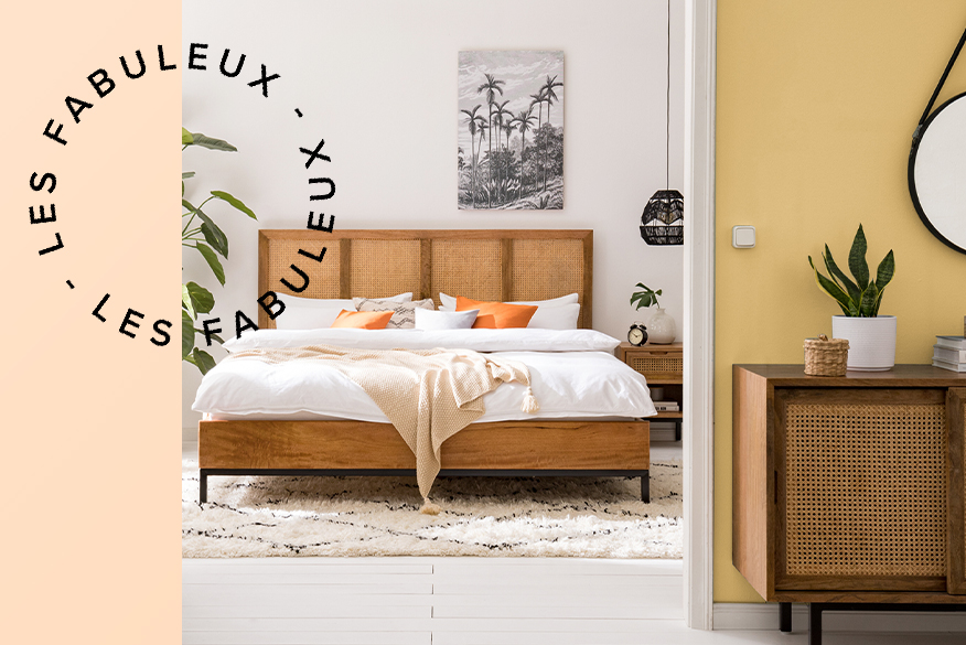 Chambre de style bohème avec mur jaune et meubles cannés, tête de lit cannée ; lit boxspring blanc avec haute tête de lit capitonnée, coussins et attrape-rêves.