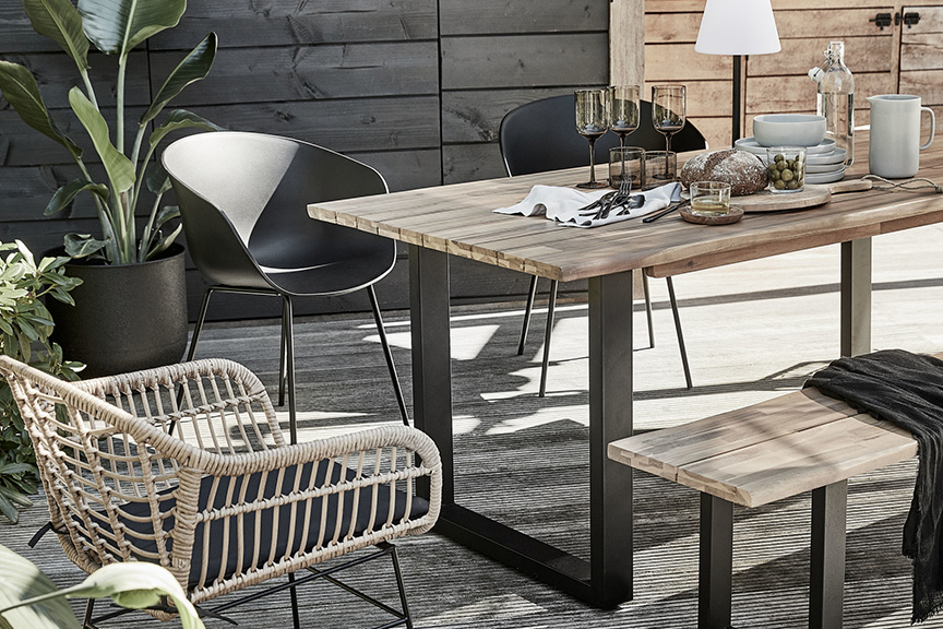 Terrasse im roughen Industrial-Style mit Outdoor-Möbeln aus Akazienholz und Eisengestell, dazu ein Rattanstuhl plus weitere Stühle mit schwarzer Sitzschale, Tisch eingedeckt samt Steingutgeschirr, dunklen Gläsern und Holzbrettchen, als Highlight eine moderne Outdoor-Akkuleuchte.