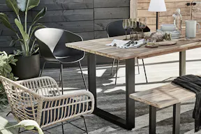 Terrasse im roughen Industrial-Style mit Outdoor-Möbeln aus Akazienholz und Eisengestell, dazu ein Rattanstuhl plus weitere Stühle mit schwarzer Sitzschale, Tisch eingedeckt samt Steingutgeschirr, dunklen Gläsern und Holzbrettchen, als Highlight eine moderne Outdoor-Akkuleuchte.