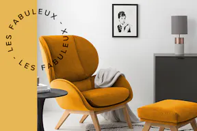 Fauteuil rétro en velours jaune orangé avec tabouret assorti sur un tapis noir et blanc, accompagné d'une table d'appoint noire, d'un buffet gris mat, d'un miroir, d'une lampe de table en béton ainsi que d'une affiche représentant Audrey Hepburn