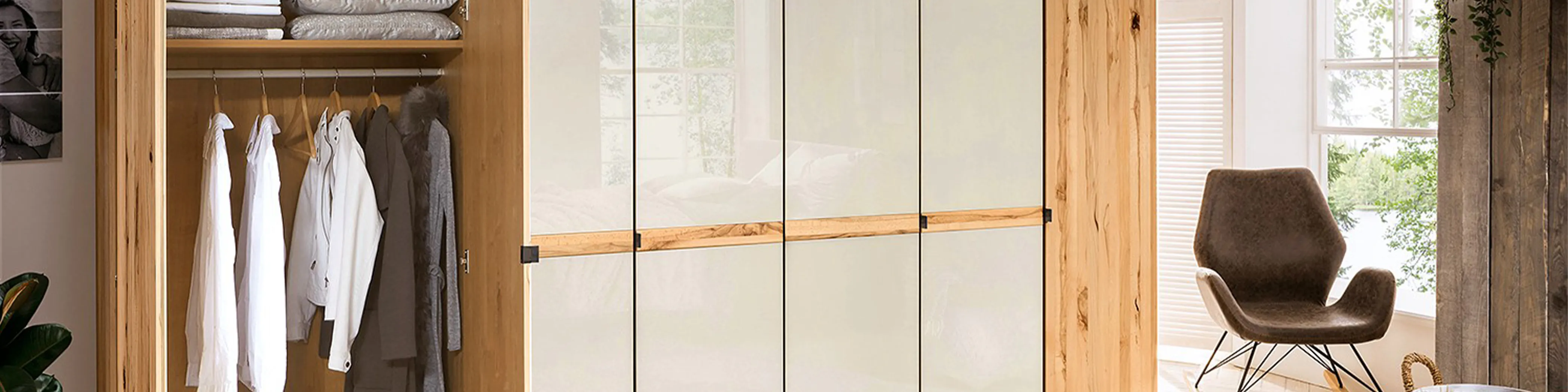 Schicker Kleiderschrank mit Holz- und Milchglastüren, eine Seite geöffnet mit Kleidung auf Bügeln, dahinter ist ein Sessel vor einem offenen Fenster