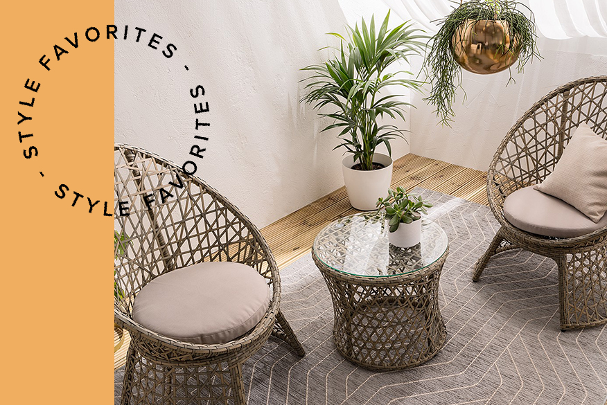 Outdoor-Rattanmöbel in einem Indoor-Wohnraum; zwei Rattansessel mit Kissen und ein runder Rattantisch mit Glasplatte, darunter ein grauer Outdoor-Teppich, ringsum viele Grünpflanzen wie Palmen und Sukkulenten
