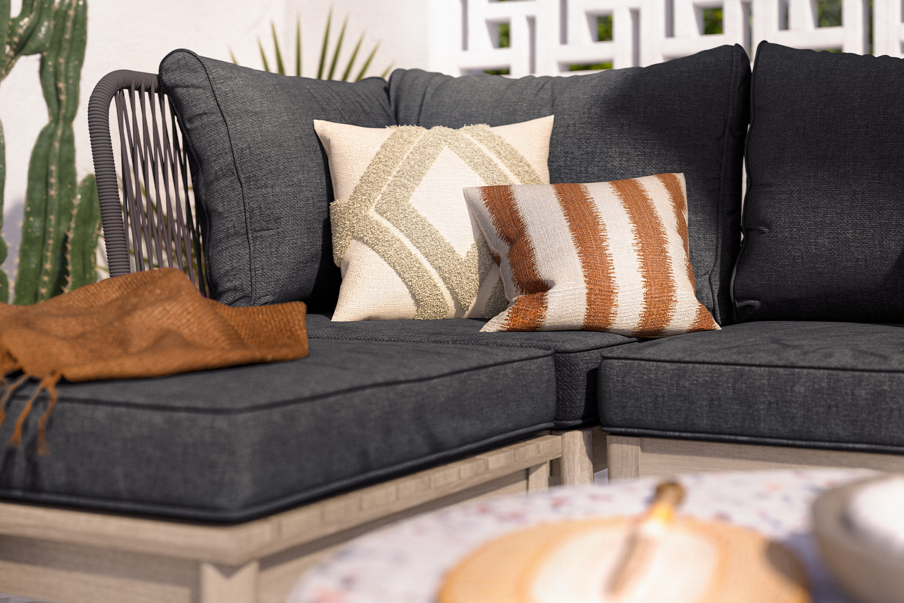 Outdoor-Loungesofa mit grauen Polstern, dazu ein beige-weiß und braun-weiß gemustertes Kissen und eine dunkelbraune Decke.