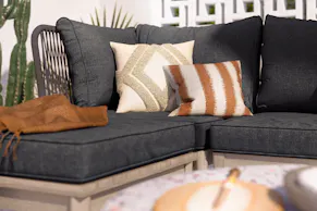 Outdoor-Loungesofa mit grauen Polstern, dazu ein beige-weiß und braun-weiß gemustertes Kissen und eine dunkelbraune Decke.