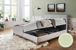 Hellgraues Polsterbett mit zwei Bettkästen für mehr Stauraum im Schlafzimmer, welches vor mintgrüner Wand auf einem Holzfußboden steht.