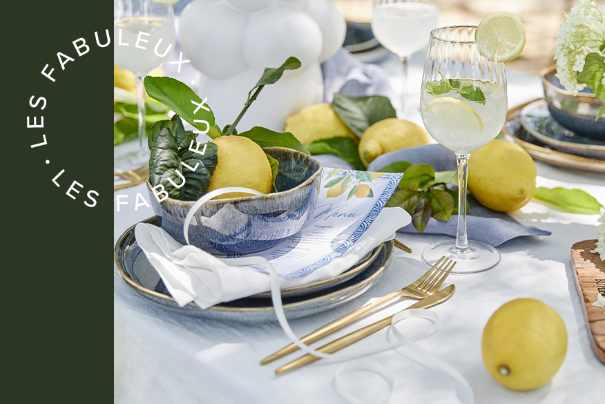 Table dressée avec du linge de table blanc, de la vaisselle bleue, des couverts dorés et de beaux citrons jaunes avec leurs feuilles pour la décoration, le tout accompagné de Limoncello Spritz dans des verres à pied BUTLERS.