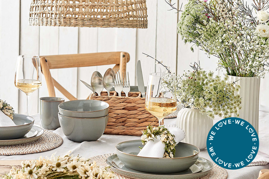 Gedeckter Holztisch mit weißem Tischläufer, blau-grauem Keramikgeschirr, Besteckkasten und Platzteller aus Rattan sowie einer Deko mit hellen Trockenblumen in der Vase als auch als Blumenkranz.