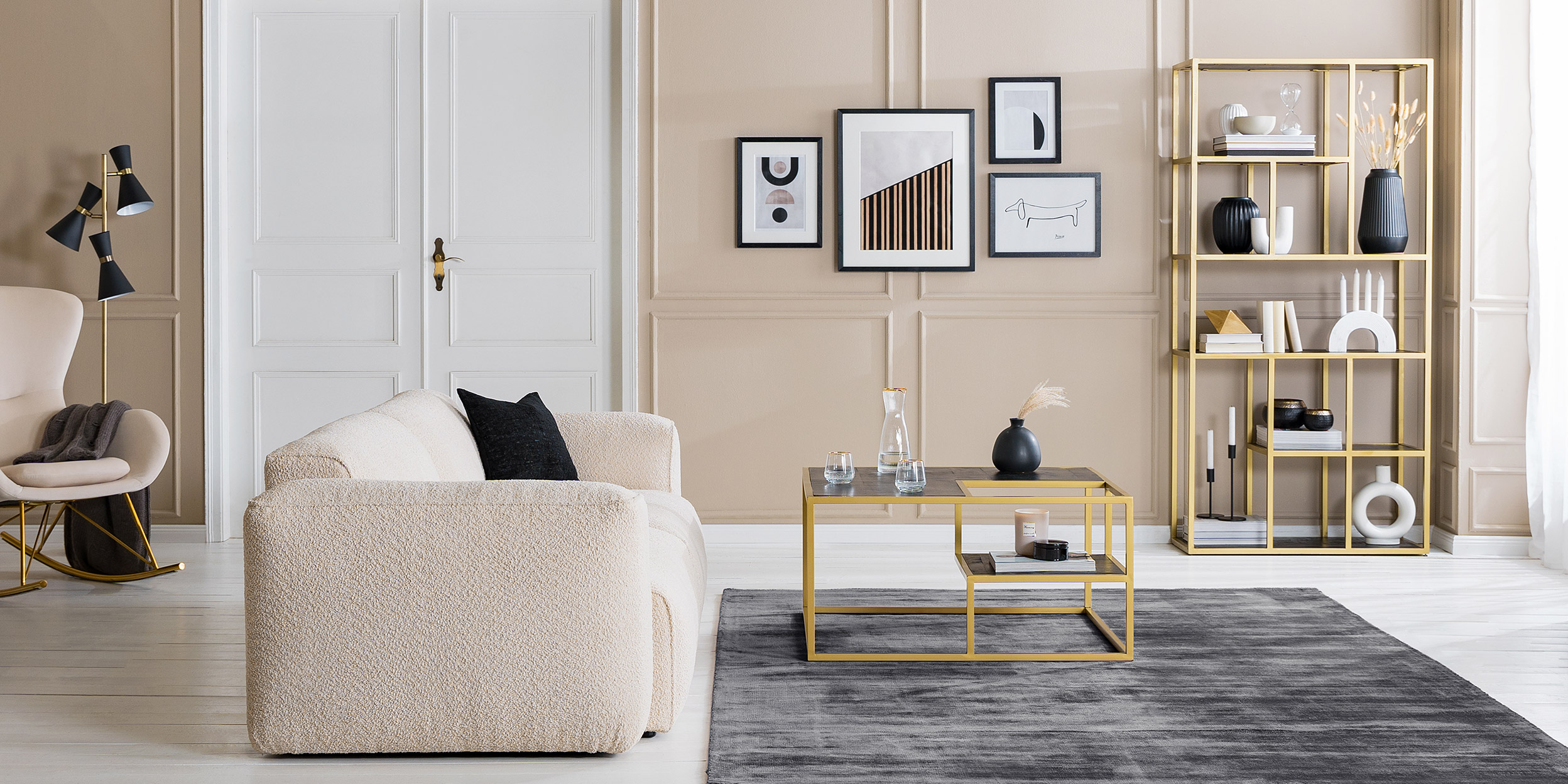 Wohnzimmer mit einem beigen Bouclésofa und -sessel; ein Couchtisch, Regal, TV-Board sowie eine Kommode in einer eleganten Schwarz-Gold-Kombination, dazu schwarze und weiße Deko und viele Textilien.