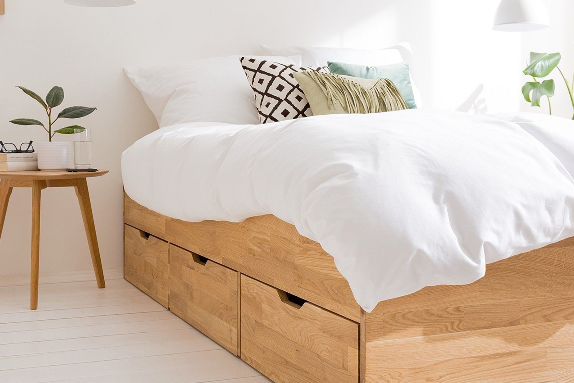Stauraumbett aus hellem Holz mit 3 Schubkästen, weißer Bettwäsche und weißen Pendellampen über dem filigranen Nachttisch.