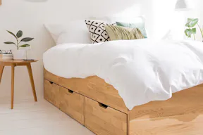 Stauraumbett aus hellem Holz mit 3 Schubkästen, weißer Bettwäsche und weißen Pendellampen über dem filigranen Nachttisch.