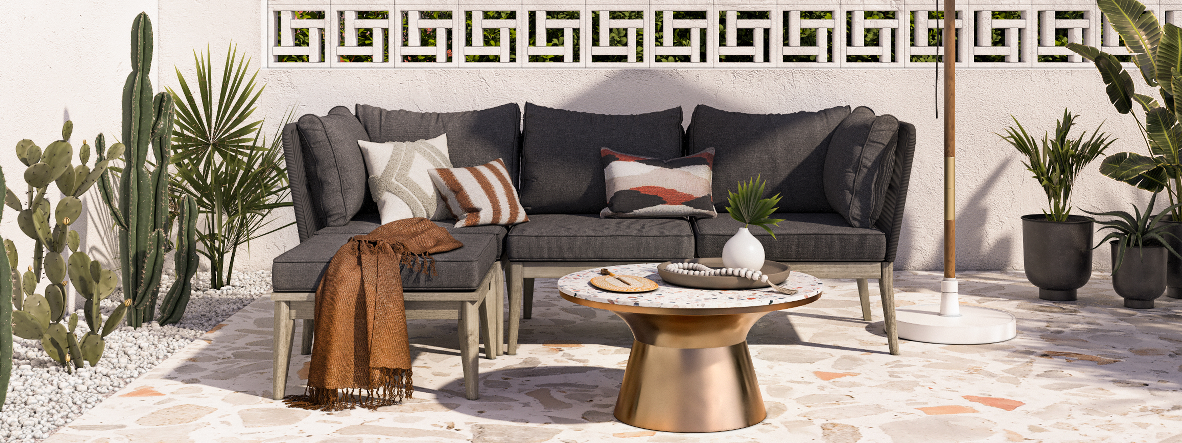 Terrasse im orientalischen Stil mit einem Loungesofa mit grauen Polstern und gemusterten Kissen in Erdtönen, dazu ein kupferfarbener Tisch mit Terrazzo-Platte, ein Sonnenschirm, Kakteen und sonstige Grünpflanzen.