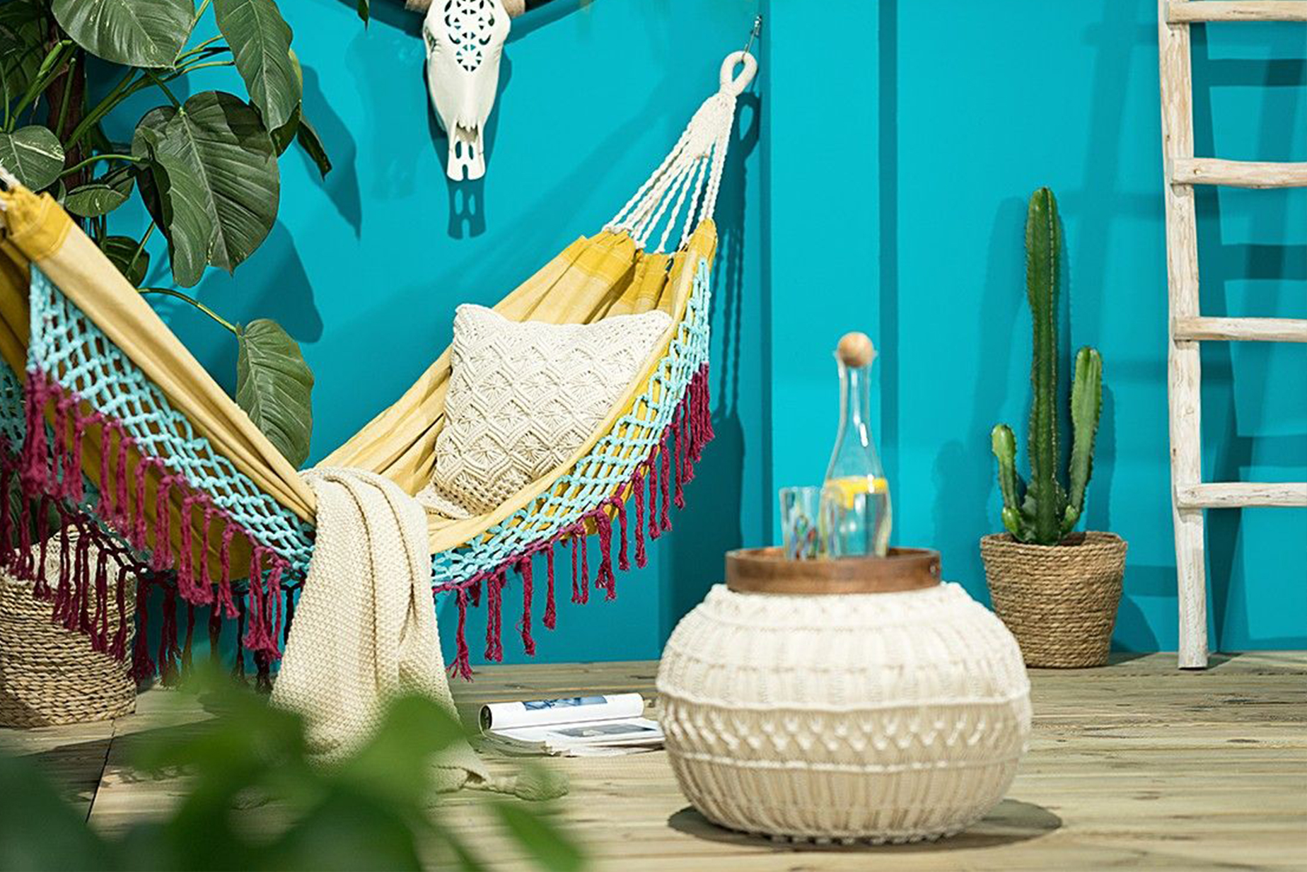 Bunte Hängematte mit Fransen, dazu eine beige Decke und Kissen mit Strickmuster, ein beiger Strickpouf, ein Deko-Geweih sowie ein Kaktus und Grünpflanzen in Blumenampeln.