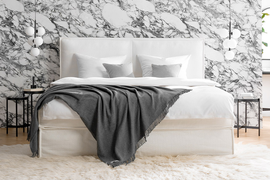 Weißes Boxspringbett KINX der home24 Exklusivmarke Studio Copenhagen vor einer Wand mit moderner Marmortapete, daneben schlanke Beistelltische, Hängelampen und ein Stuhl für abgelegte Kleidung mit Wiener Geflecht.