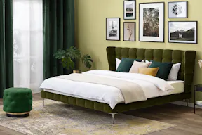 Schlafzimmer im Hotelstil mit einem grünen Polsterbett aus Samt mit weißer Bettwäsche, beigem Plaid und mehreren Kissen in Dunkelgrün, Beige und Gold. Vor der lindgrünen Wand steht ein runder, schwarzer Nachttisch mit Büchern und goldenem Kerzenständer; darüber hängt eine goldene Pendelleuchte aus Glas.