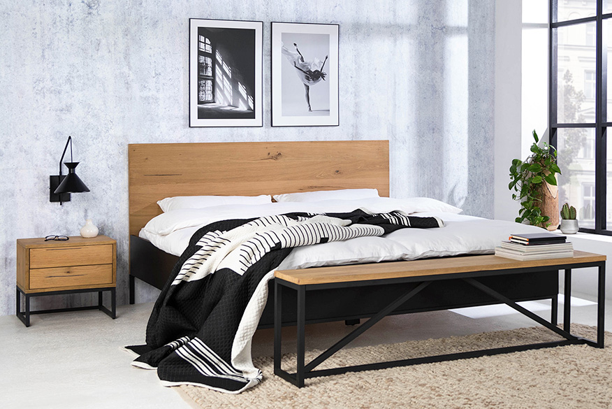 Schlafzimmer im minimalistischen Stil mit Bett, Nachttisch und Sitzbank der Serie Flox im Industrial-Stil aus Eichenholz und schwarzem Metall. Dazu schwarz-weiße Wandbilder, eine schwarze Wandleuchte, ein beiger Teppich und ein schwarzer Garderobenständer aus Metall.