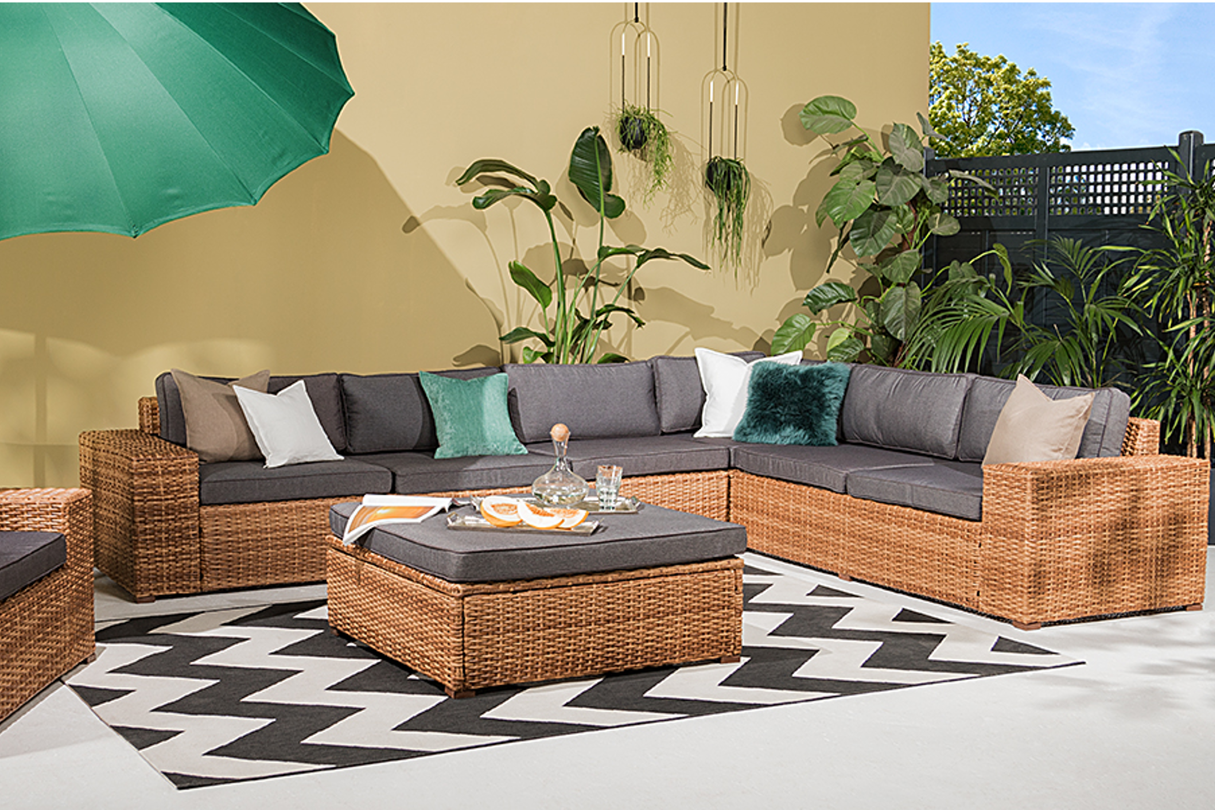 Geräumige Gartenlounge in heller Rattanoptik mit grauen Polstern, umrahmt von Pflanzen