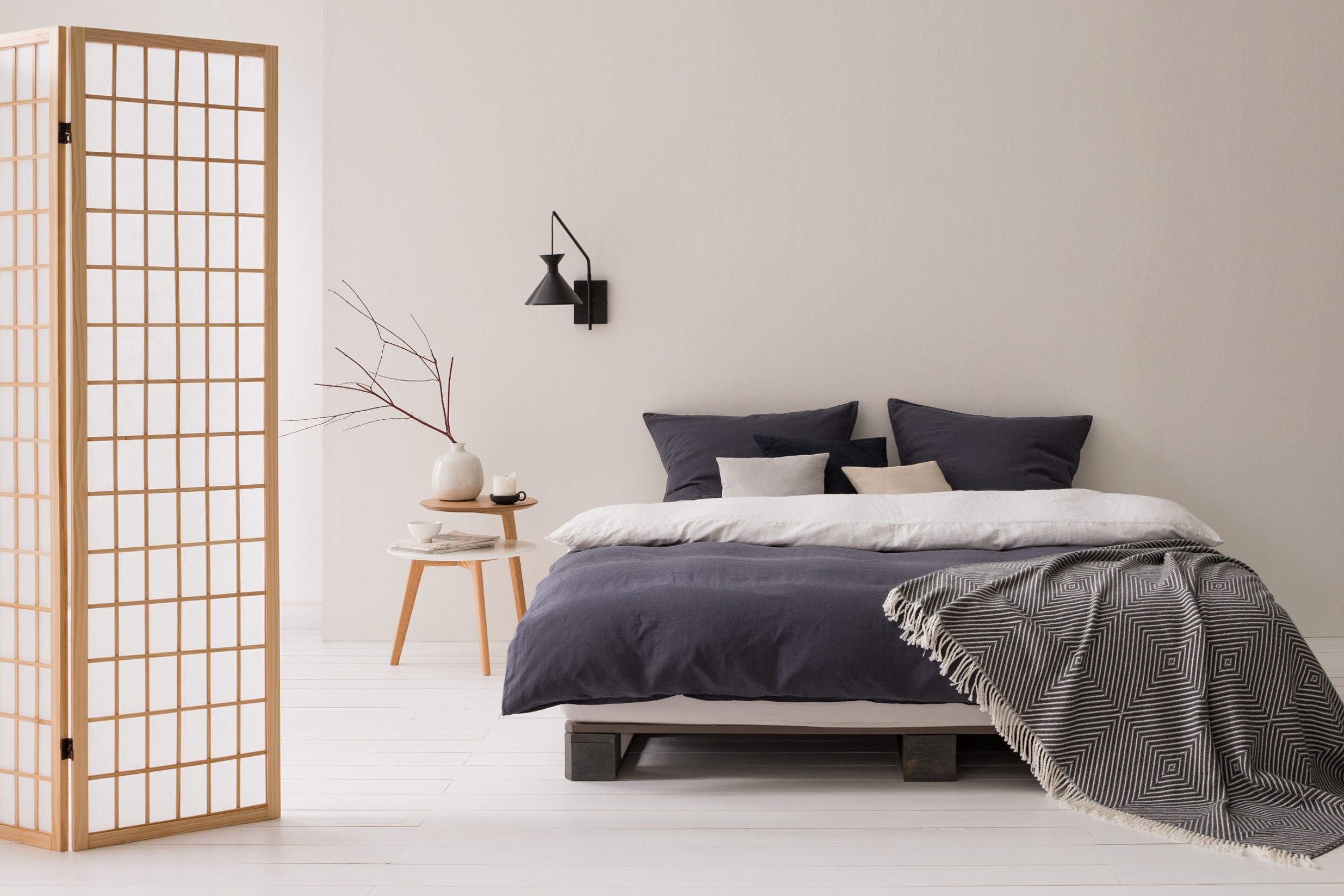 Schlafraum mit Wandlampe, runden Tischen, einfachem Bett und Paravent im Vordergrund