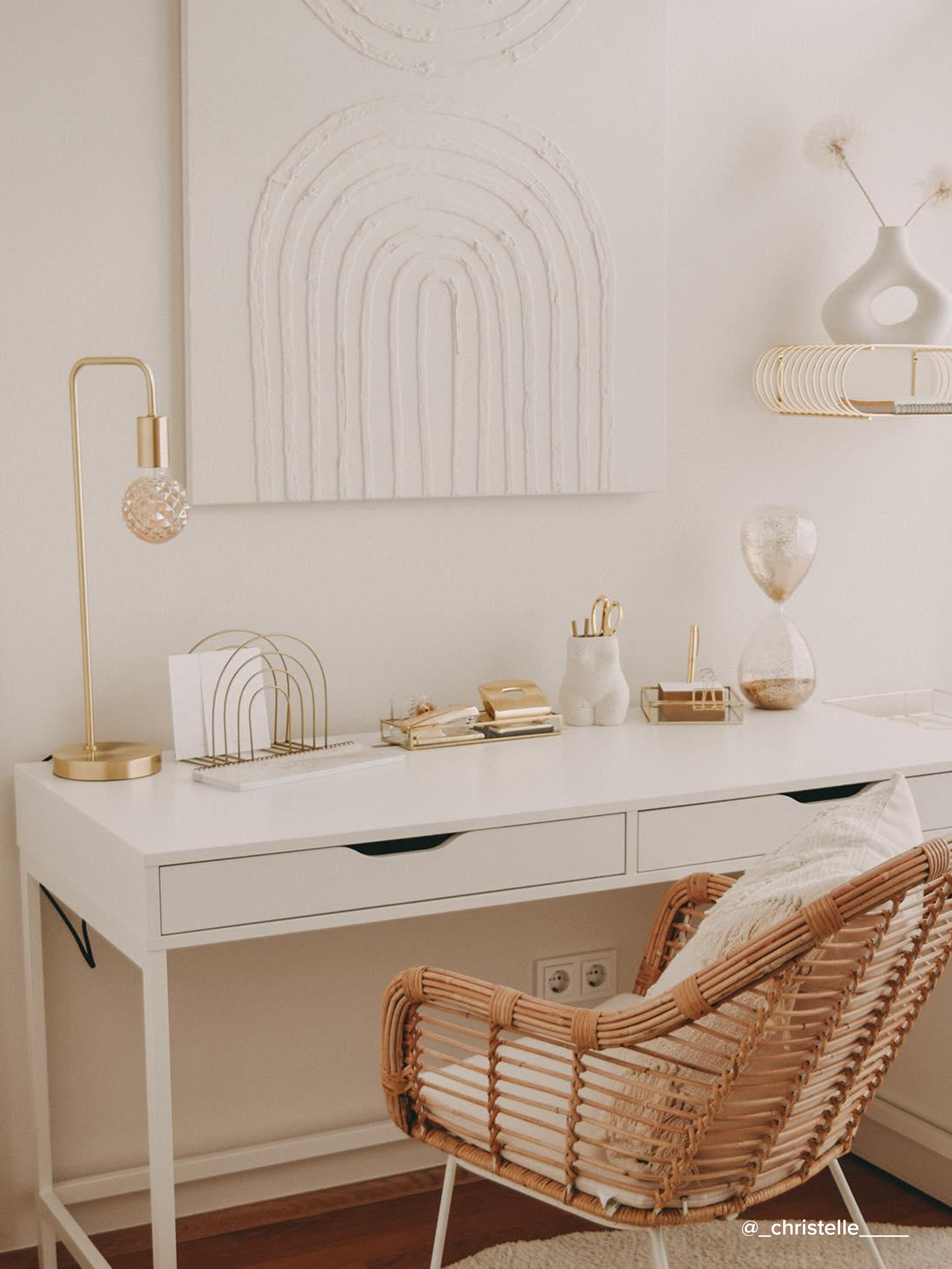 Praktisch eingerichtetes Homeoffice mit einem weißen Schreibtisch, einem Rattanstuhl im Boho-Stil, goldenen Wohnaccessoires sowie einem weißen Strukturbild.