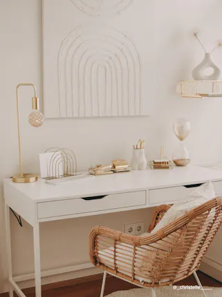 Praktisch eingerichtetes Homeoffice mit einem weißen Schreibtisch, einem Rattanstuhl im Boho-Stil, goldenen Wohnaccessoires sowie einem weißen Strukturbild.