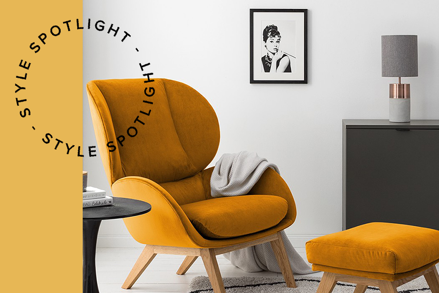 Retro fauteuil van oranjegeel fluweel met bijpassende hocker, op een zwart-wit vloerkleed, plus een zwart bijzettafeltje, een matgrijs dressoir, een decoratieve spiegel, een betonnen tafellamp en een afbeelding van Audrey Hepburn; ernaast een zwarte metalen hanglamp.