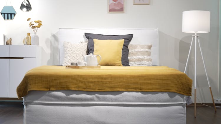 Boxspringbett KINX in Weiß gezeigt in einem der home24 Showrooms, dekoriert mit Kissen, Stehlampe und ebenfalls weißem Sideboard.