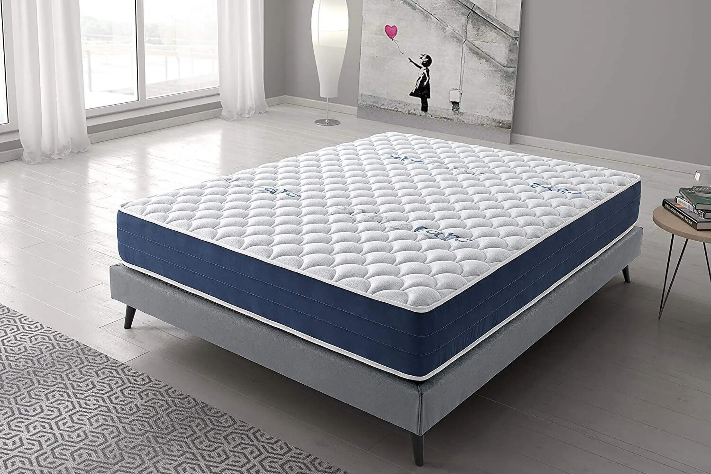 Einfaches Bettgestell in Grau mit dicker Matratze in einem kahlen Raum mit einem Banksy-Kunstdruck