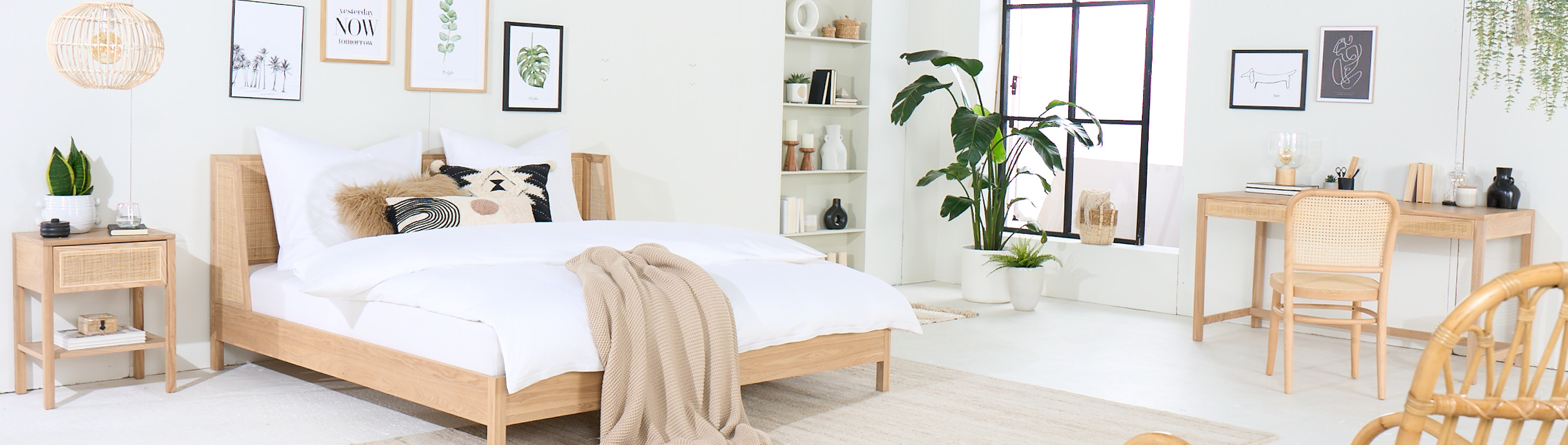 Schlafzimmer mit hellen Holzmöbeln der Serie Taylor by kollected, einer Exklusivmarke von home24: ein Bett, ein Nachttisch, ein Schaukelstuhl, ein Schreibtisch inkl. Schreibtischstuhl, dazu viele Grünpflanzen, Teppiche, Textilien und ein Strickpouf. Allen Taylor-Produkten gemein ist die Kombination aus hellem Eichenholz und Rattan.