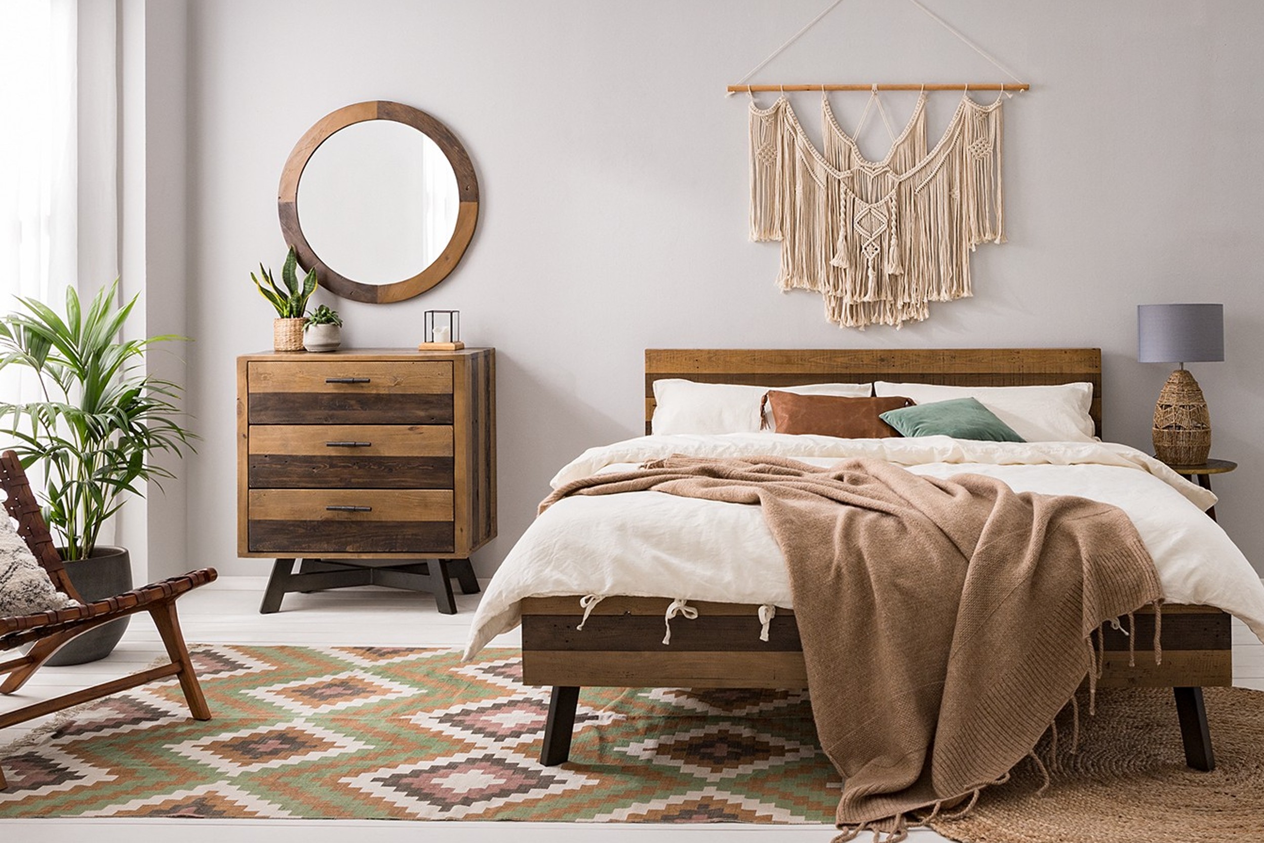 Chambre à coucher aux murs blancs, avec un tapis à motifs tons nature (vert, marron), un lit et des meubles en bois, une literie blanche, des rideaux blancs, et des éléments décoratifs en bois.