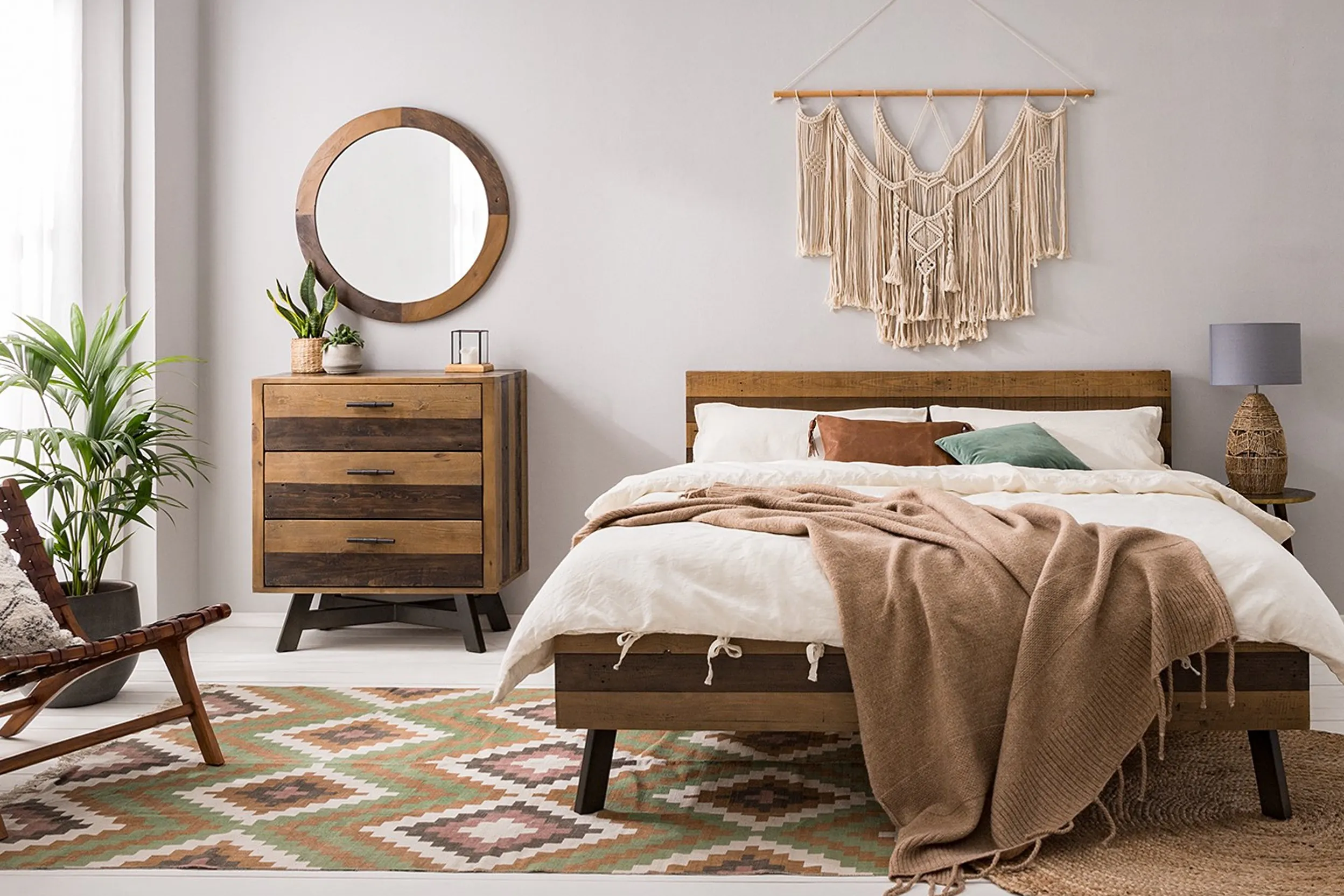 Chambre à coucher aux murs blancs, avec un tapis à motifs tons nature (vert, marron), un lit et des meubles en bois, une literie blanche, des rideaux blancs, et des éléments décoratifs en bois.
