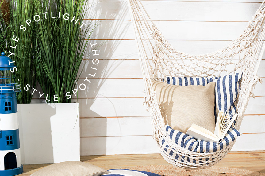 Witte hangfauteuil met blauw-wit gestreept zitkussen en beige kussens voor een witte houten muur; daarnaast een foto van een helderblauwe lucht met witte wolken.