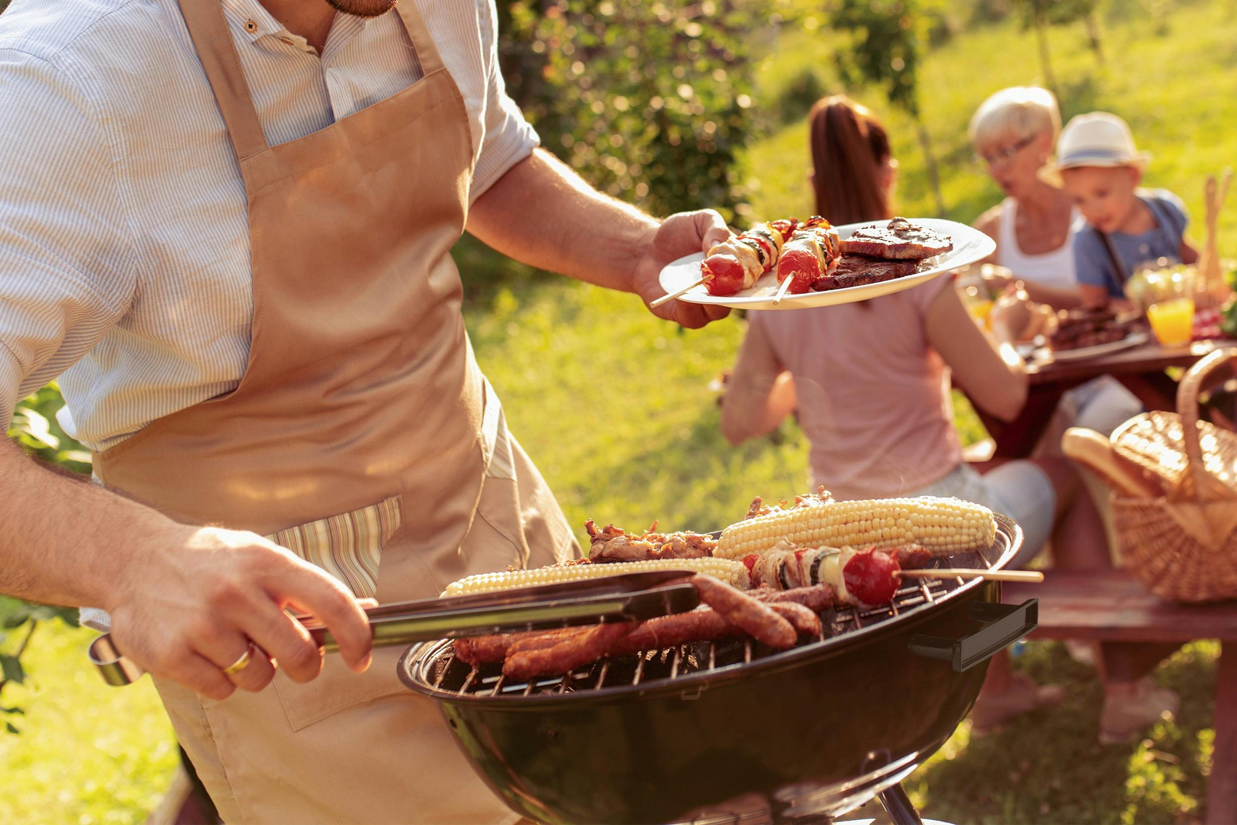 Ein Mann mit Schürze steht an einem Kugelgrill, während im Hintergrund einige Leute essen.