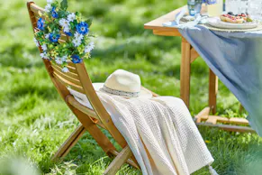 Ein Holzgartenstuhl von BUTLERS mit einer cremeweissen Sommerdecke vor einem Gartentisch mit Picknick-Equipment.