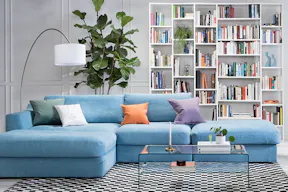 Wohnzimmer mit hellblauem Sofa, bunten Kissen, einem Glastisch sowie einem grünem Samtsessel, dazu eine Bogenlampe mit weißem Schirm, ein weißes Regal und ein schwarz-weiß karierter Teppich.