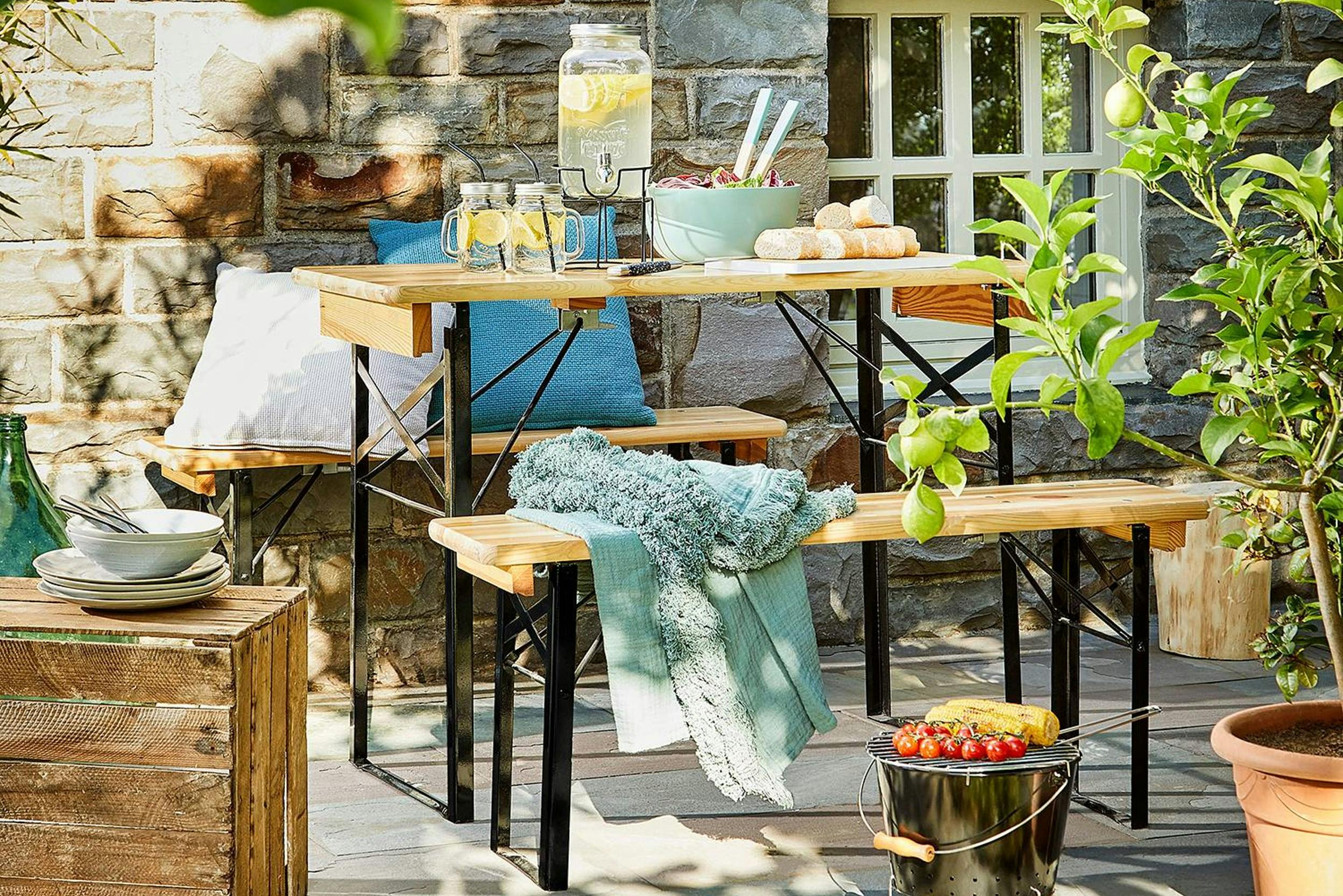 Bierzeltgarnitur mit Getränkespender, Limogläser, Salat und Brot hinter einem Eimergrill