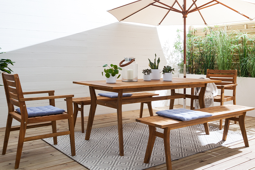 Terrasse mit Gartentisch, Gartenstühlen und Gartenbank aus Akazienholz mit hellblauen Sitzkissen, dazu ein weißer Sonnenschirm, ein gemusterter Outdoor-Teppich und Deko.