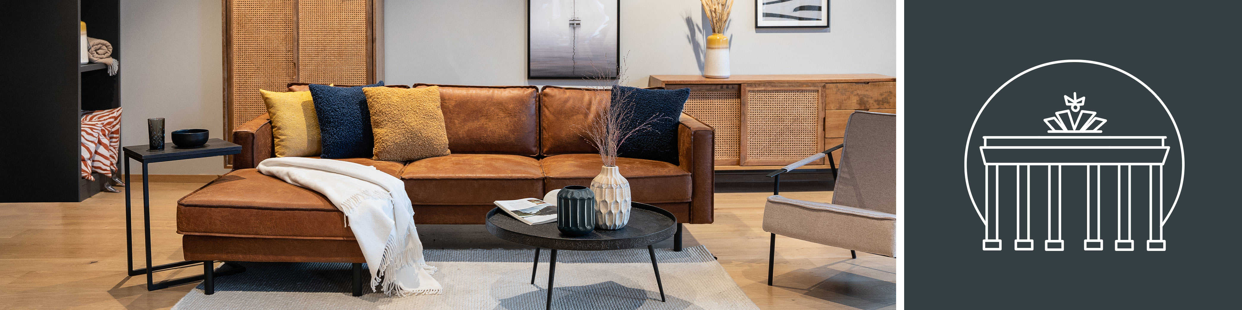 Ecksofa Fort Dodge der home24 Exklusivmarke kollected in der Farbe Cognac im home24 Showroom Berlin Prenzlauer Berg, dazu ein jeweils schwarzer Couch- und Beistelltisch, ein grauer Armlehnenstuhl, zwei Vasen, eine weiße Decke, ein grauer Teppich sowie ein Sideboard und Highboard mit Wiener Geflecht der Serie Vina by kollected; daneben das Brandenburger Tor als Symbol für den home24 Showroom in Berlin