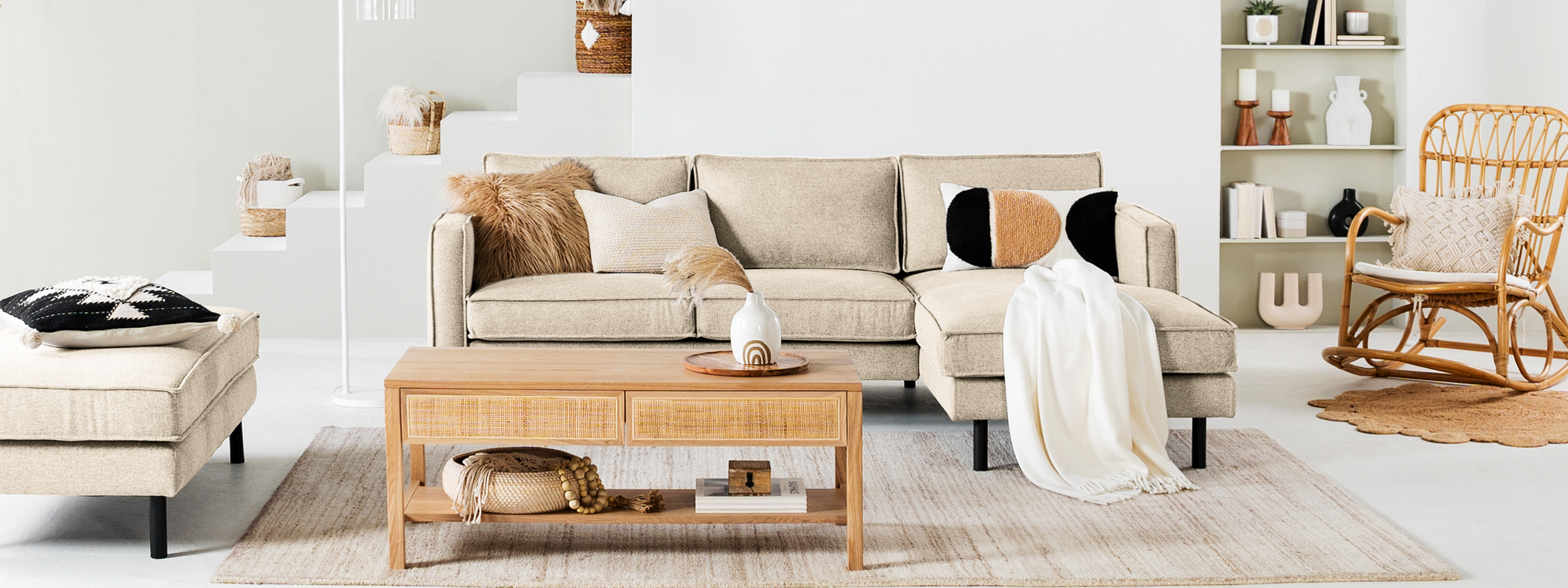 Wohnzimmer mit Möbeln der Serien Fort Dodge und Taylor der home24 Exklusivmarke kollected: Im Mittelpunkt ein beiges Sofa, dazu ein Couchtisch mit geflochtenen Applikationen, Pendelleuchten aus Rattan, Kissen, Teppich und Plaid in Beige- und Cremetönen sowie viele Dekoobjekte im Wandregal.
