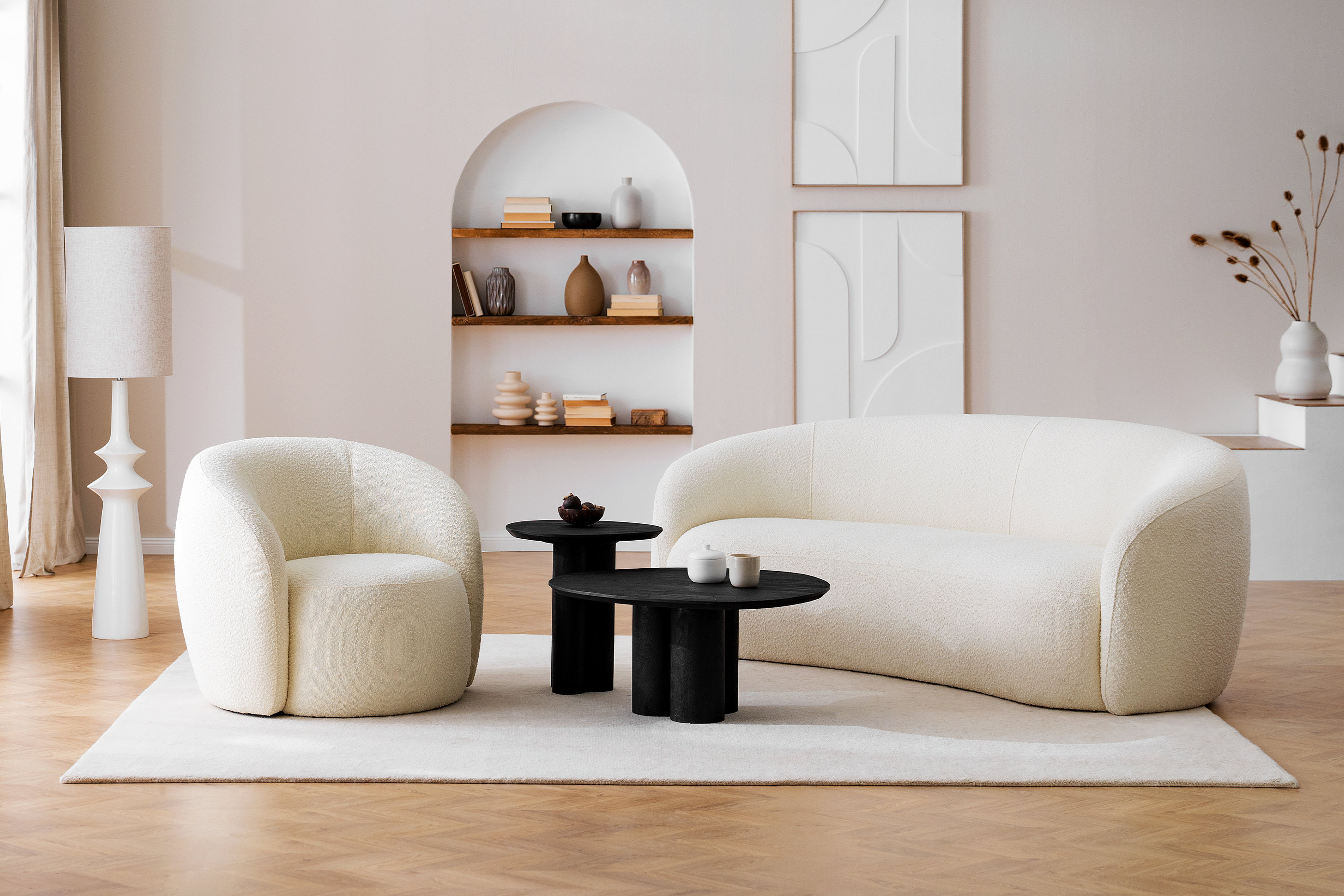 Wohnzimmer in organischen Formen mit Sessel und Sofa Osane in Creme sowie dem schwarzen Couchtisch Besare von Studio Copenhagen, dazu ein weißer Teppich, eine weiße Steh- und Tischleuchte, Keramik- und Holzdeko in Erdtönen sowie ein schwarzes Sideboard.
