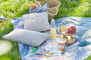 Eine blau-gemusterte Picknickdecke von BUTLERS mit weissen Kissen, dazu ein Picknickkorb sowie Gläser und eine Etagere.