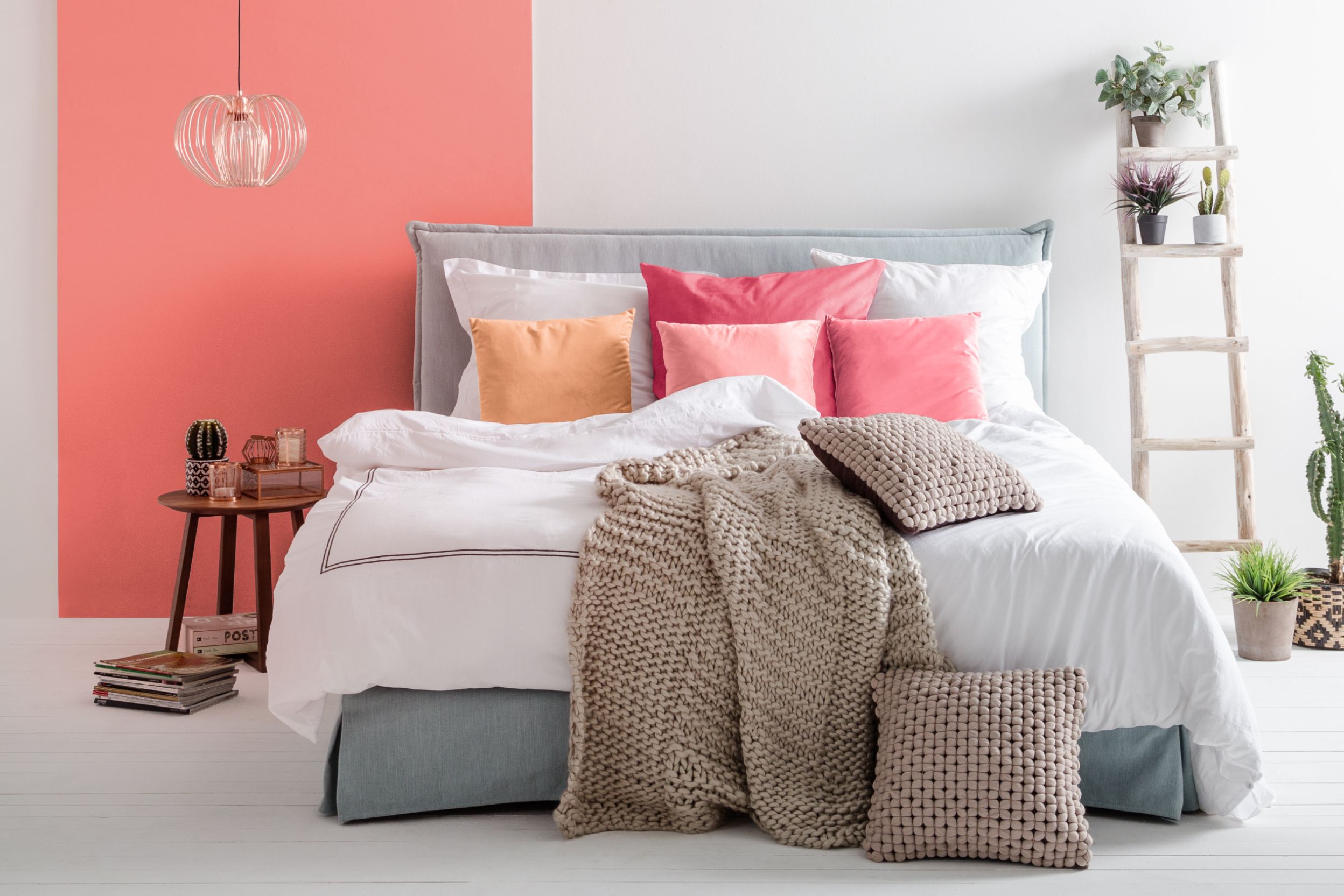 Chambre à coucher dont le mur est rose/corail et gris clair, et dont le lit est gris clair, blanc, avec des coussins dans les tons roses. roses