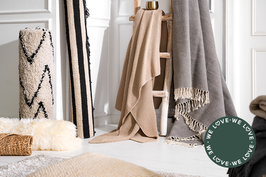 Textilien wie Teppiche und Decken aus verschiedenen kuschelig-flauschen Materialien wie Wolle, Faux-Fur, Grobstrick und Fransen für eine gemütliche Winterzeit, daneben weihnachtliche Dekoration auf einem Schafsfell.