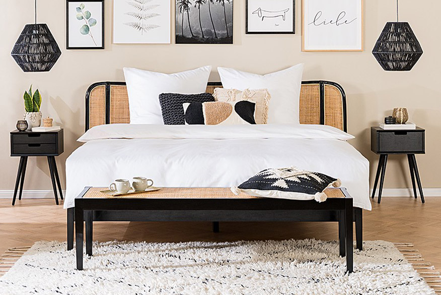 Schlafzimmer im Retro-Look mit Bett, Sideboard, Schreibtisch und Stuhl mit Rattangeflecht im Gittermuster der home24 Exklusivmarke kollected. Dazu eine Bilderwand mit verschiedenen Motiven, ein Dekospiegel, zwei schwarze Hängelampen aus Rattan sowie helle Textilien und Pflanzen.