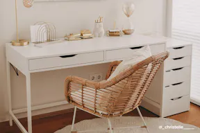 Einfach und effektvoll eingerichtetes Homeoffice mit einem weißen Schreibtisch als Basis, dazu ein Rattanstuhl im Boho-Stil und reichlich goldene Wohnaccessoire für einen Touch Glamour, darüber ein weißes Strukturbild.