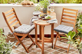 Kleiner Balkon mit Klappmöbeln aus Holz, bei dem der kleine Tisch mit Tomate-Mozzarella, Brot und Wein gedeckt ist; Grünpflanzen, graue Sitzkissen und ein beiger Outdoorteppich von BUTLERS machen's gemütlich.