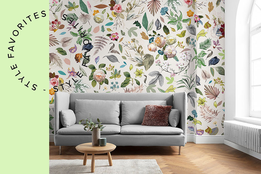 Graues Sofa vor einer Wand mit Blumenprinttapete, dazu ein runder Couchtisch aus Holz, Tischdeko sowie ein grauer Teppich; daneben schöne Blumenvasen in Grüntönen.