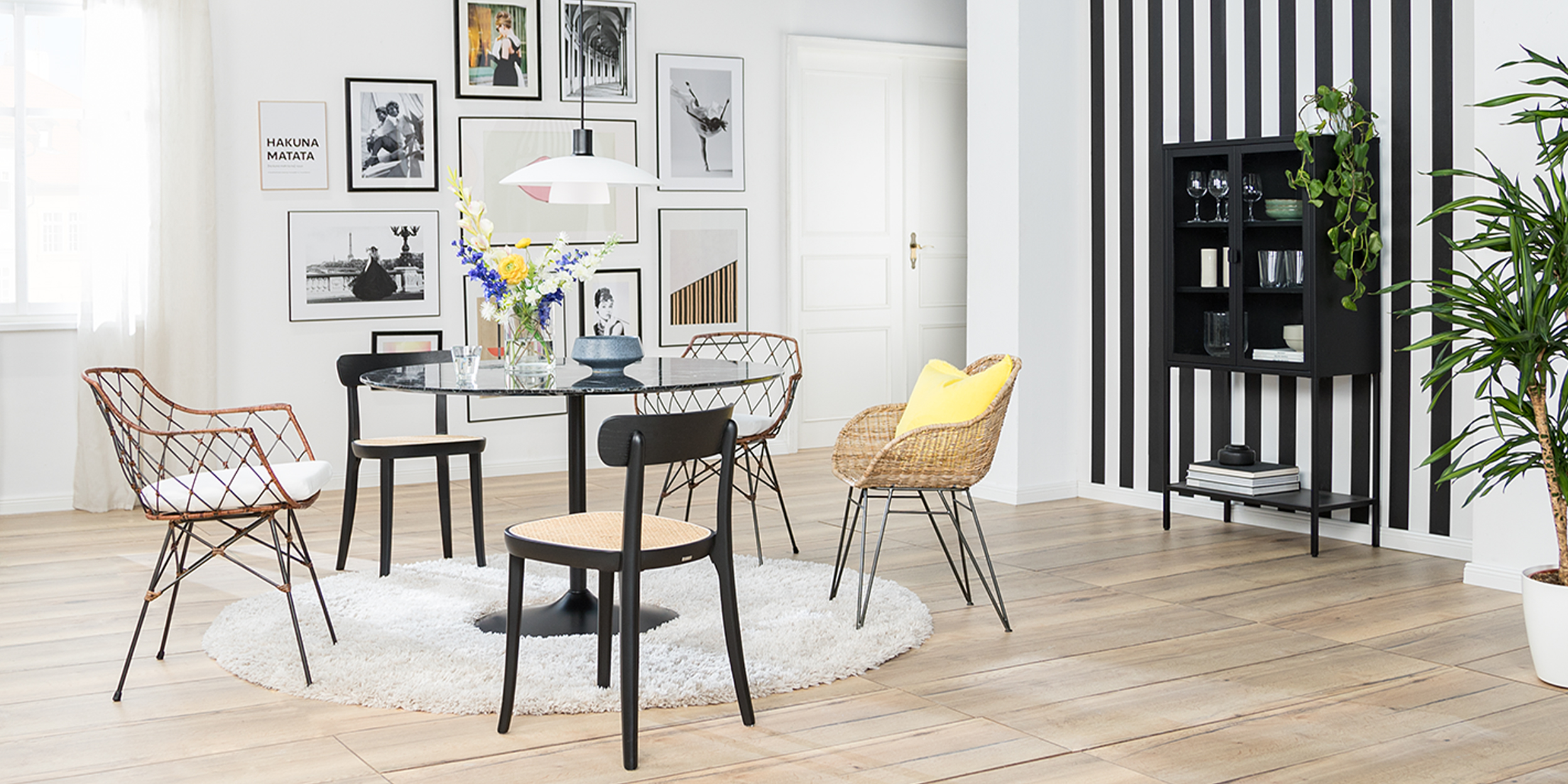 Esszimmer mit Rattan- und Holzstühlen sowie einem Glastisch in Marmoroptik. Dazu ein weißer Teppich, eine weiße Pendelleuchte, eine Bilderwand sowie ein schwarzes Highboard aus Metall vor einer schwarz-weißen Streifentapete.